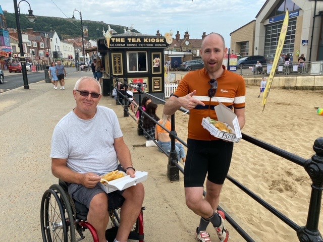 My father in a wheelchair with me looking ridiculous in cycling lycra next to him, by the beach. We’re both enjoying fish and chips, and I’m pointing at them to show it was the main reason for the ride.