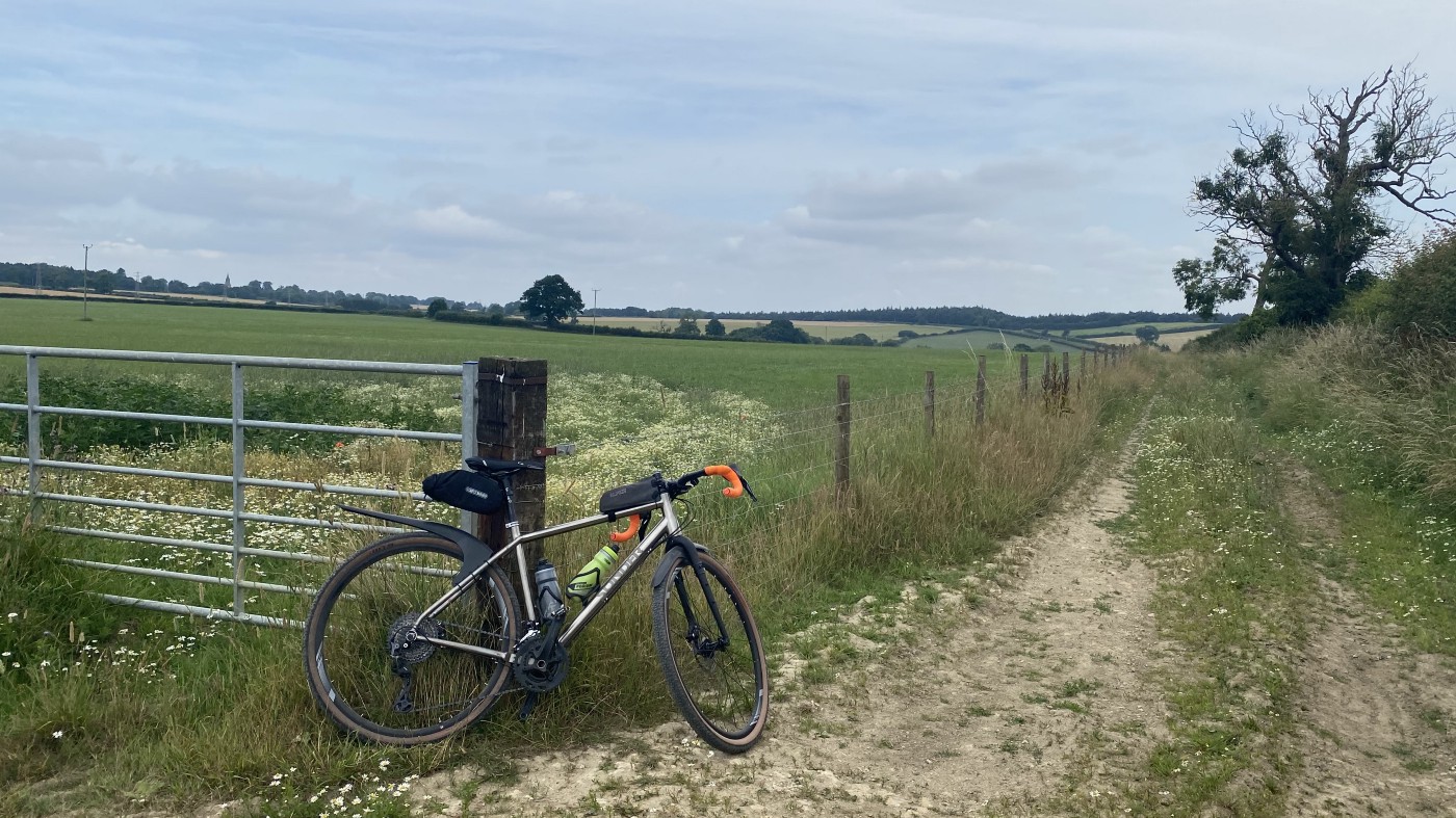 A gravel bike propped against a fencepost with a dry mud track to the right, rolling hills in the background, and slightly cloudy blue skies